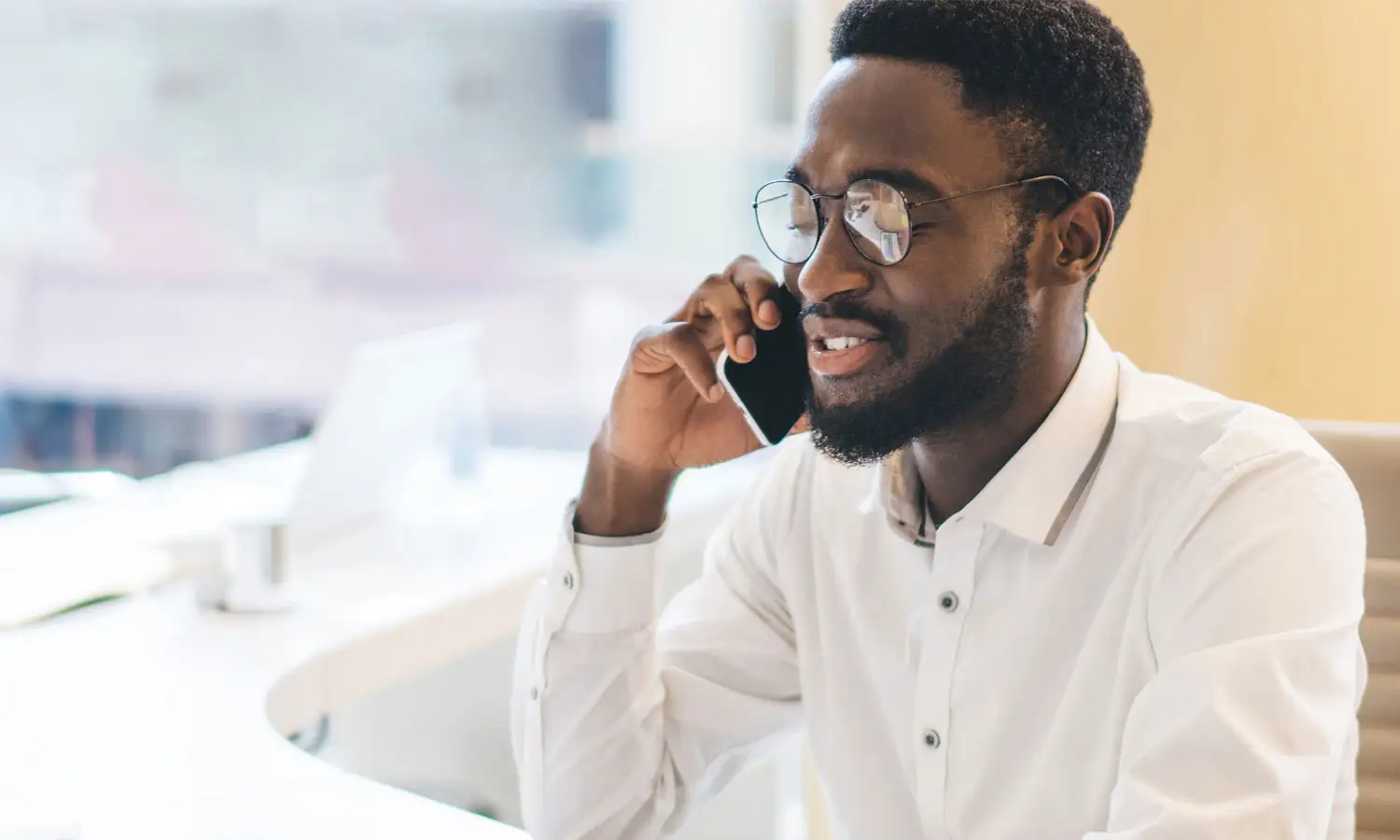 Smiling Black man with beard and glasses talking on a phone in an office.